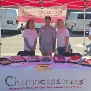Three people smiling under a pink canopy at the Chemocessories booth during the Manhattan Beach Hometown Fair. The table displays colorful accessories and informational materials, with a banner reading “Chemocessories: Warming Hearts, One Accessory at a Time.”
